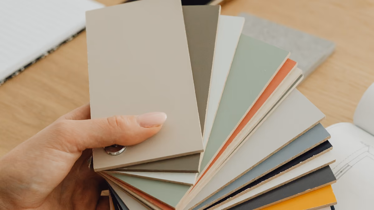 A hand holding a fanned stack of colour sample cards above a wooden desk, blueprints faintly visible behind.