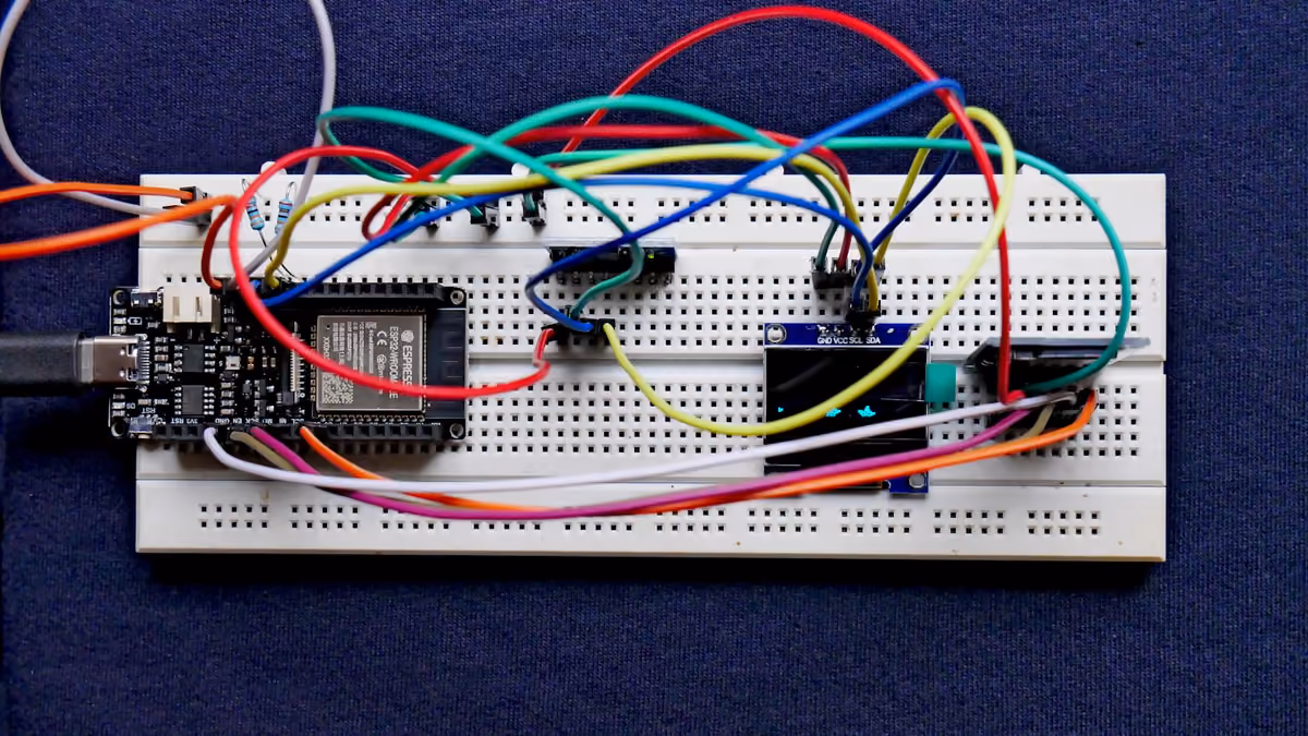 A breadboard prototype with an ESP32 board, coloured jumper wires and a small OLED display, on a blue fabric background.