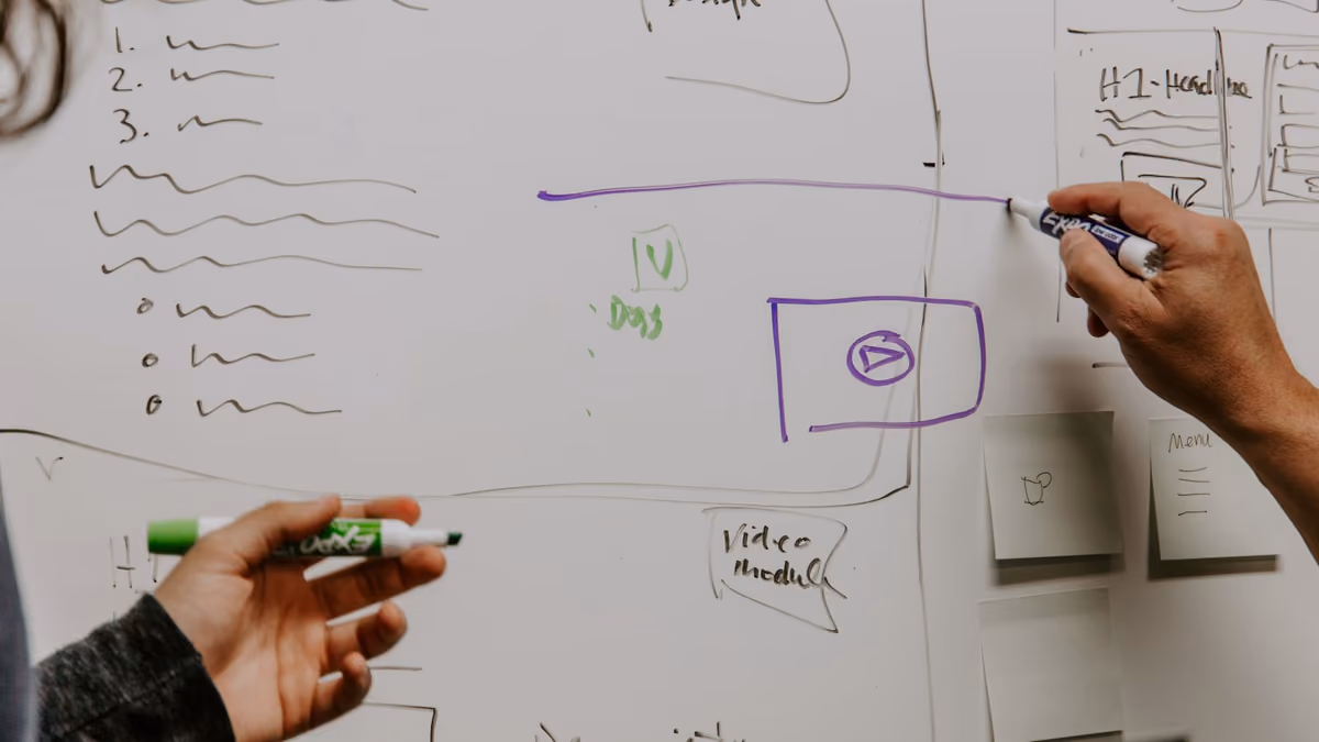 Two hands holding whiteboard markers, drawing wireframe boxes and notes beside sticky notes on a whiteboard.