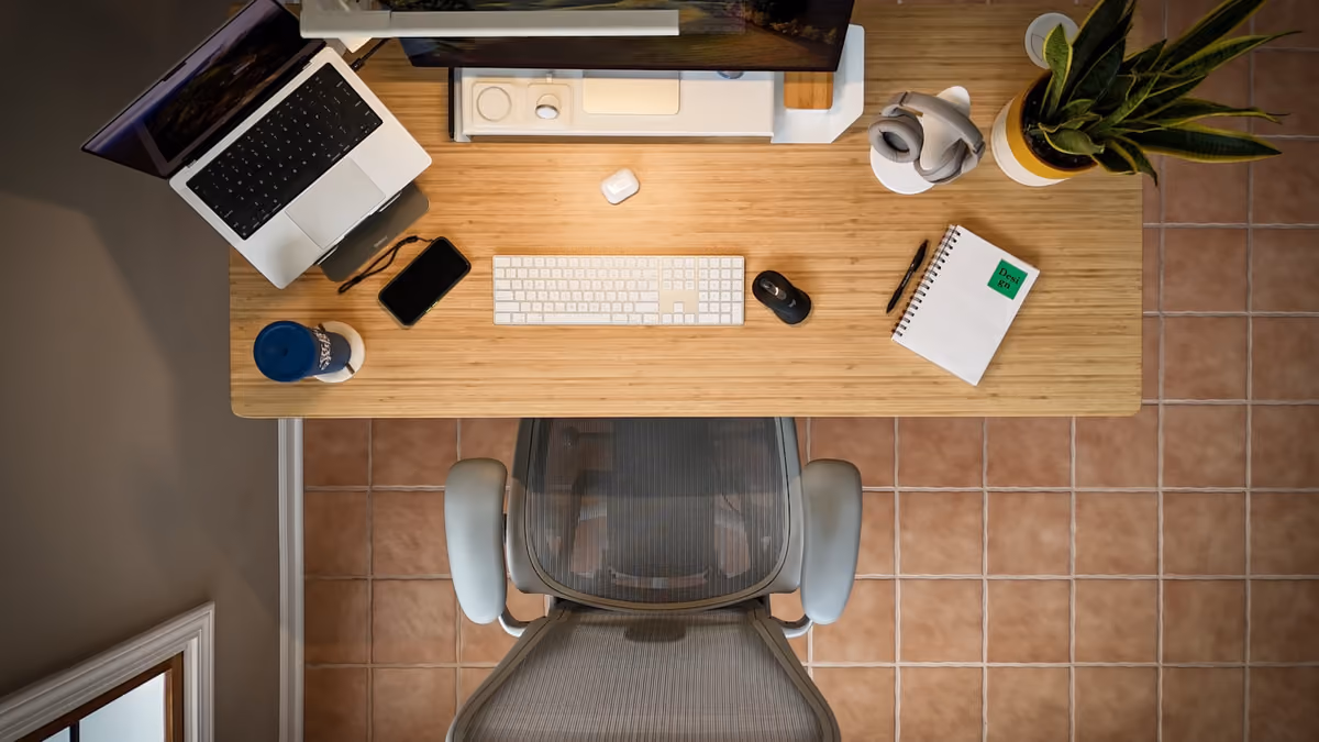 Overhead view of a desk with a laptop, keyboard, mouse, notebook, phone and a plant, empty chair beneath