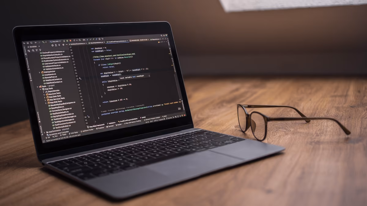 A laptop on a wooden desk showing a code editor, with a pair of glasses resting beside it